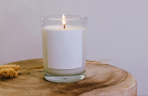 A burning, squat candle with a blank label sitting on a tree platter and plants